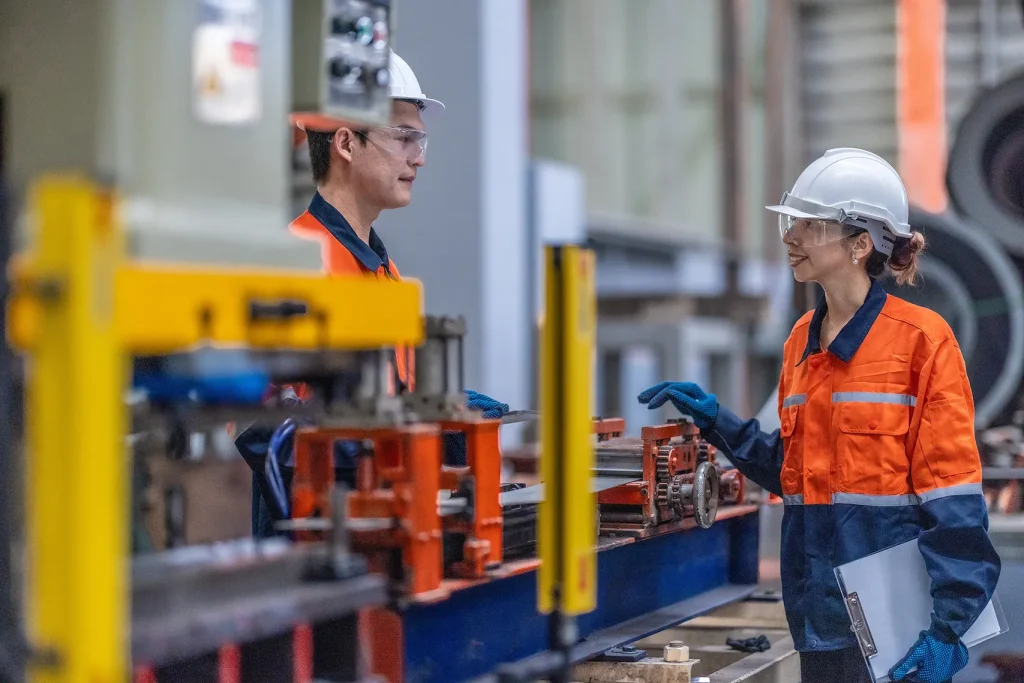 Zwei Mitarbeitende mit Schutzhelmen, Schutzbrillen und orangefarbener Arbeitskleidung an einer Produktionsmaschine.
