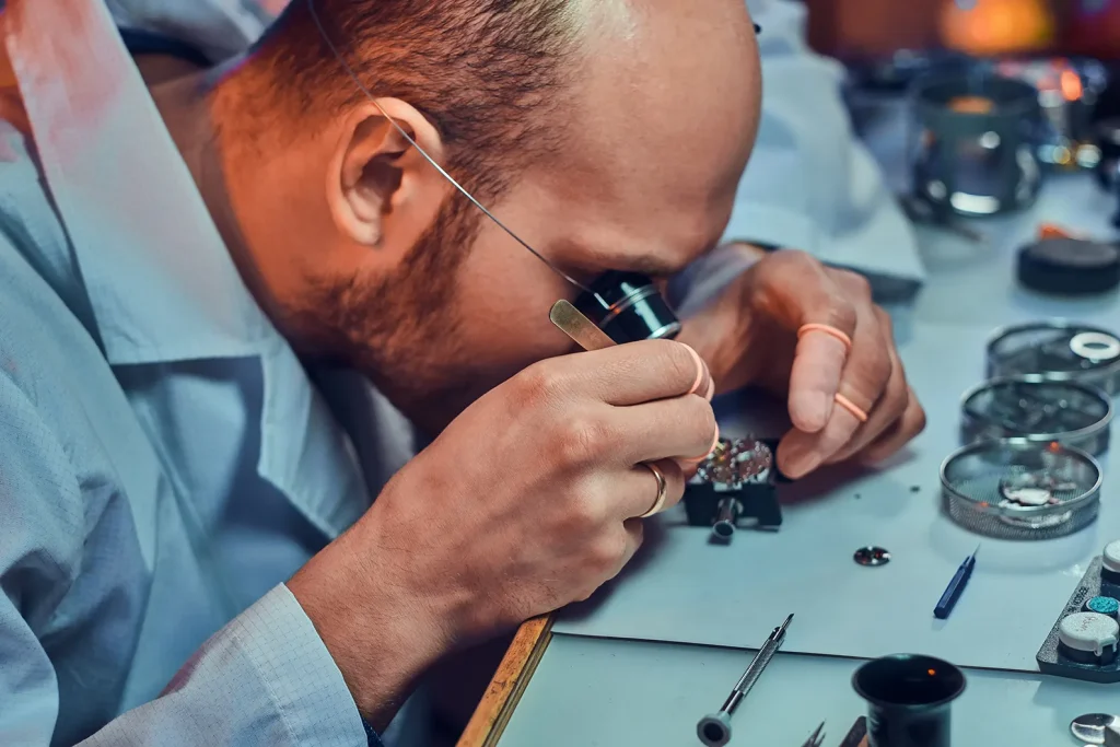 Uhrmacher bearbeitet ein Uhrwerk mit einer Augenlupe und feinem Werkzeug am Arbeitstisch.